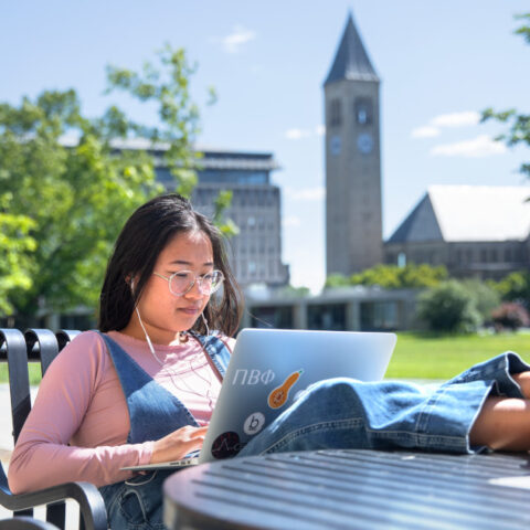 Woman sits at an outdoor table with her feet up working at a laptop, Cornell's McGraw tower and Arts Quad in the background